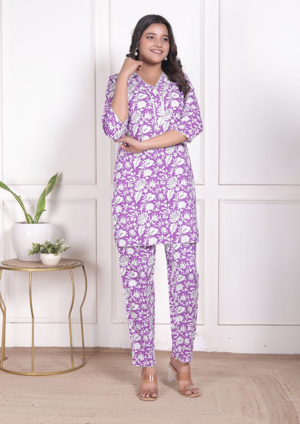 Woman wearing a purple floral pajama set in a room with a white wall and small table with plants.