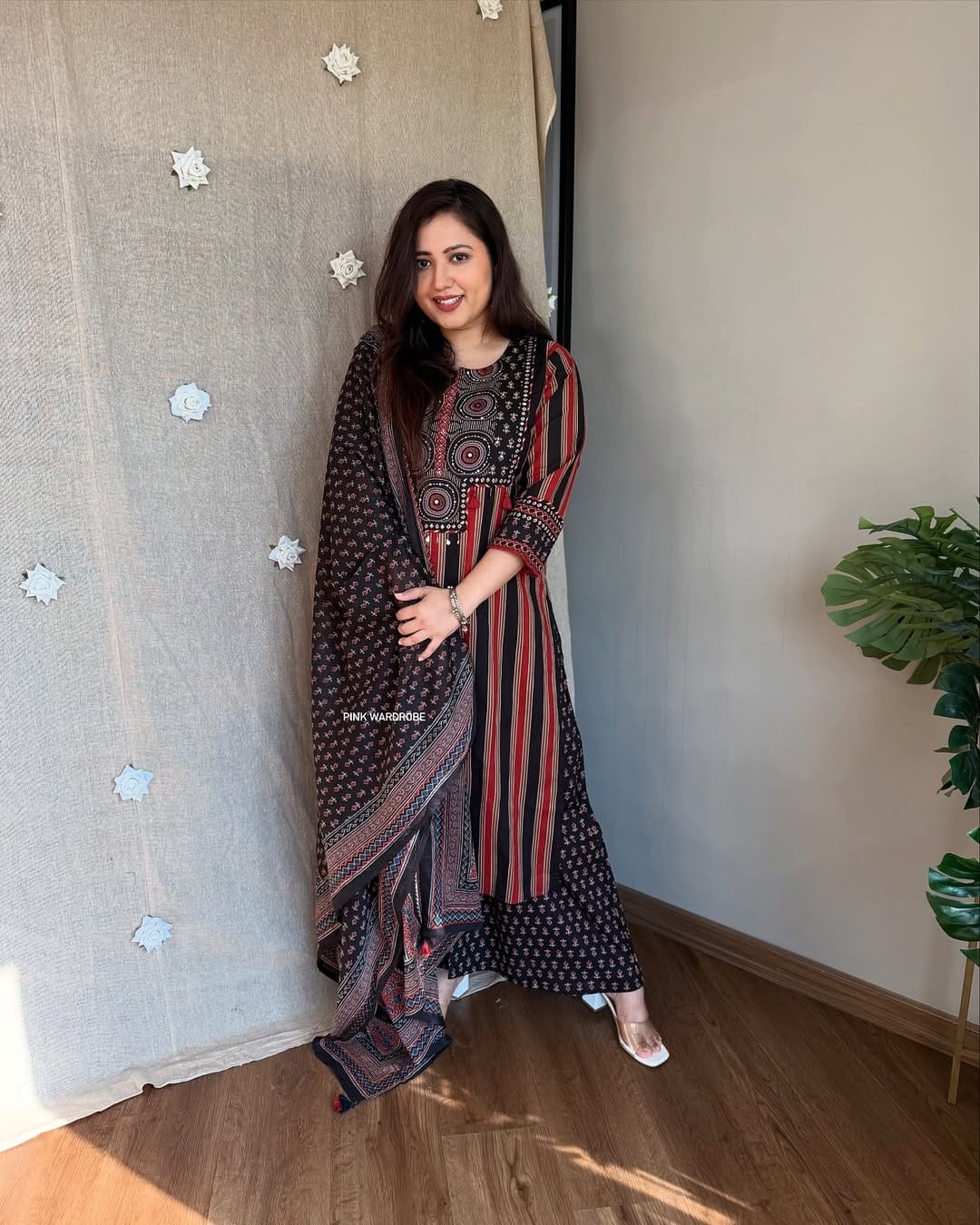 Woman in a patterned dress standing indoors with a plant in the corner.