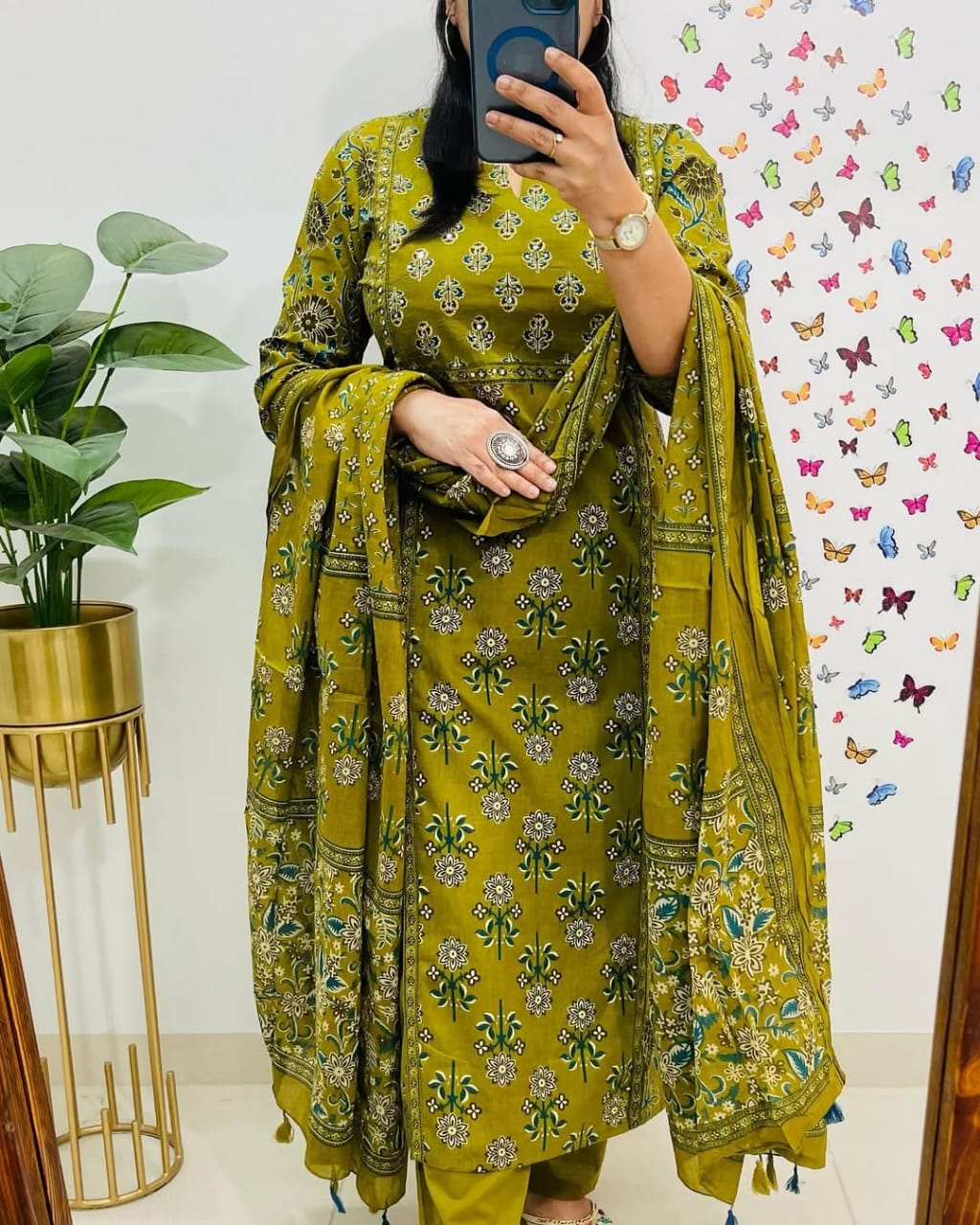 Person wearing a green floral traditional outfit with a plant and butterfly wallpaper in the background