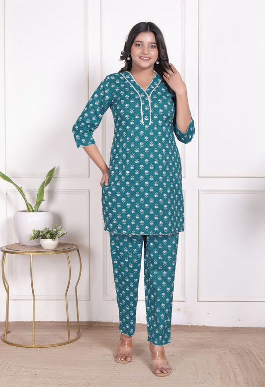 Woman wearing a teal patterned outfit standing in a room with a white wall and small table.